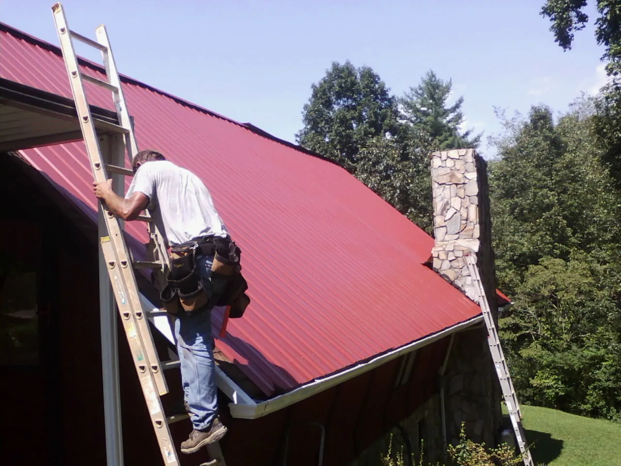 Roofer on a ladder beside a red metal roof and stone chimney — Soffit & Fascia Repair by Roofing Brothers in Wellston